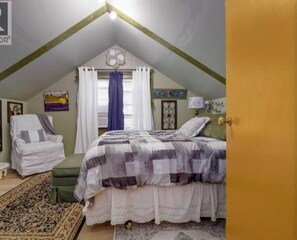 Une chambre avec un plafond en pente, un lit recouvert d’une courtepointe à carreaux, une chaise, une petite table et une fenêtre avec des rideaux.