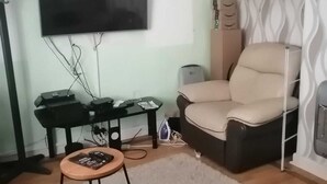 A living room with a flat-screen TV mounted on the wall, a black entertainment unit with electronic devices, a beige sofa, a small wooden stool, and a patterned curtain.
