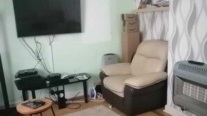 A living room with a flat-screen TV mounted on the wall, a sofa, a chair, a small round table, and a heater.
