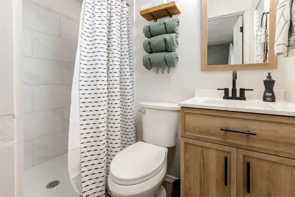 A bathroom with a white toilet, a shower with a black and white striped shower curtain, a wooden vanity with a mirror, and a towel rack with green towels.