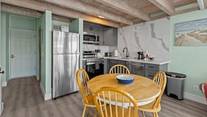 A modern kitchen with a stainless steel refrigerator, microwave, and stove. There is a dining table with four chairs and a blue bowl on it. The room has wooden beams on the ceiling and a painting on the wall. - Corner View at Cedar Cove! Gulf View Downtown! (Cedar Key)