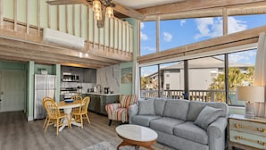 A spacious living area with a gray sectional sofa, wooden dining table, and chairs. A kitchen with modern appliances is visible in the background. - Corner View at Cedar Cove! Gulf View Downtown! (Cedar Key)