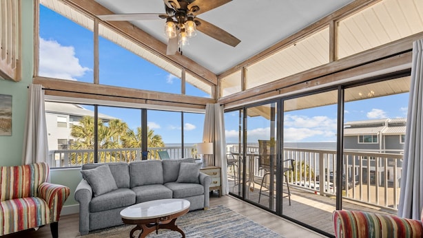 A spacious balcony with a gray sofa, striped armchairs, and a small table, overlooking a coastal area with palm trees and a clear sky. - Corner View at Cedar Cove! Gulf View Downtown! (Cedar Key)