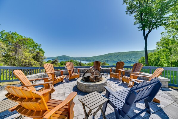 An outdoor deck with Adirondack chairs, a fire pit, and a scenic view of a lake and hills. - Finger Lakes Vacation Rental w/ Hot Tub & Pool (Naples)