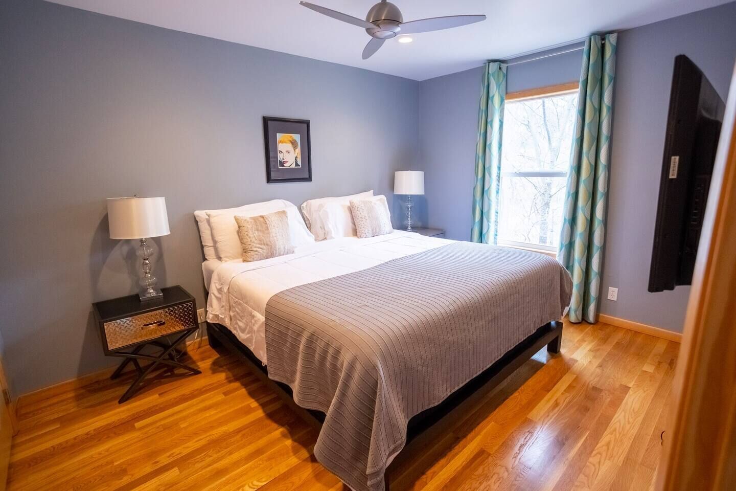 Une chambre à coucher avec un lit, deux lampes de chevet, un ventilateur de plafond, un tableau encadré au mur et une fenêtre avec des rideaux.
