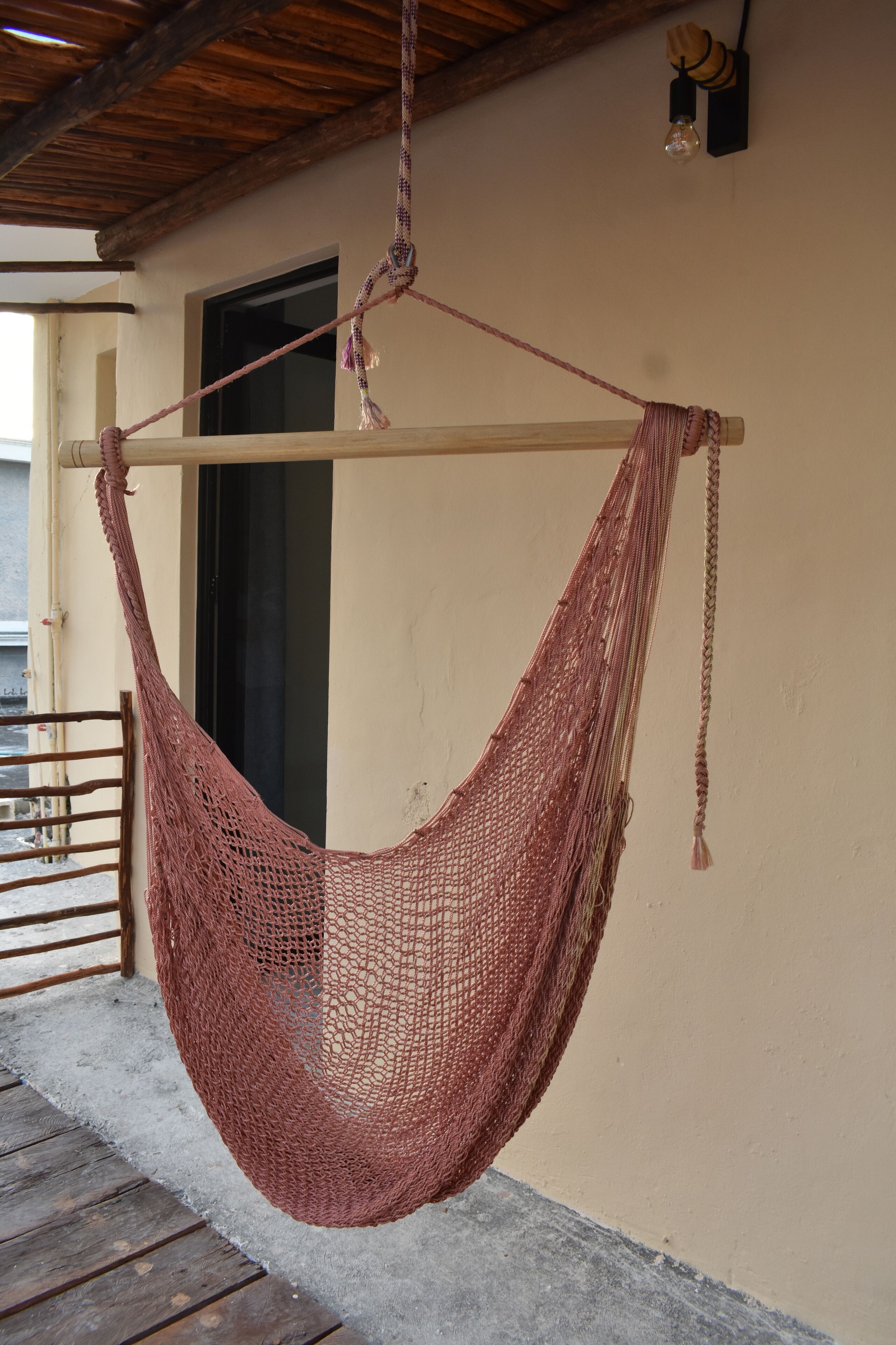 Una hamaca colgada de una viga de madera, sujeta a una pared color beige con una ventana y una puerta.