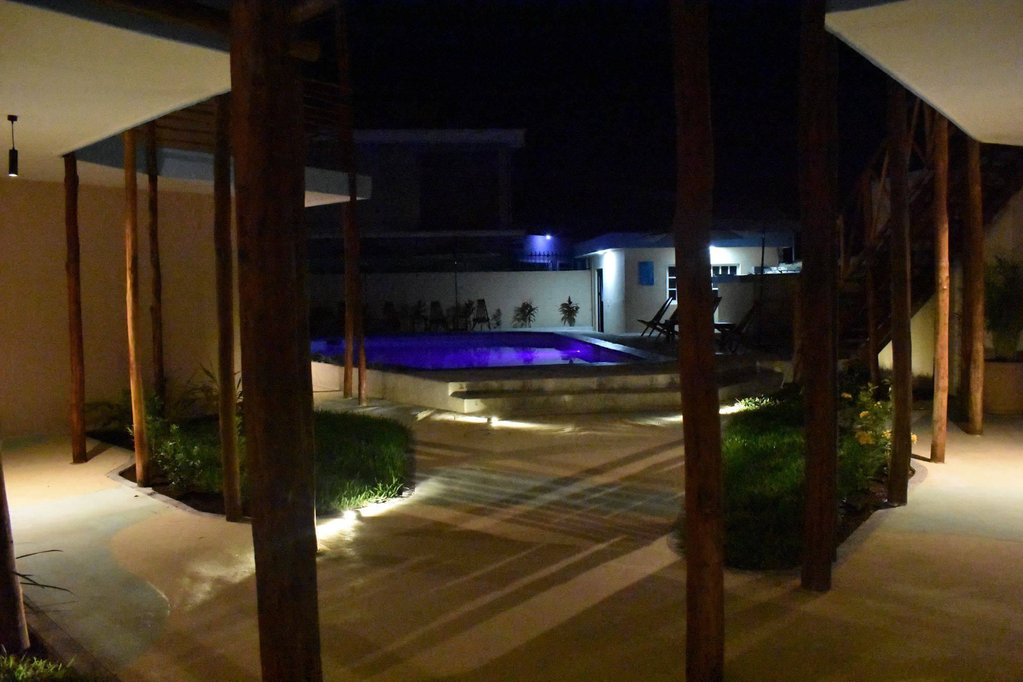 A nighttime view of a pool area with a blue-lit pool, surrounded by a covered patio with wooden beams.
