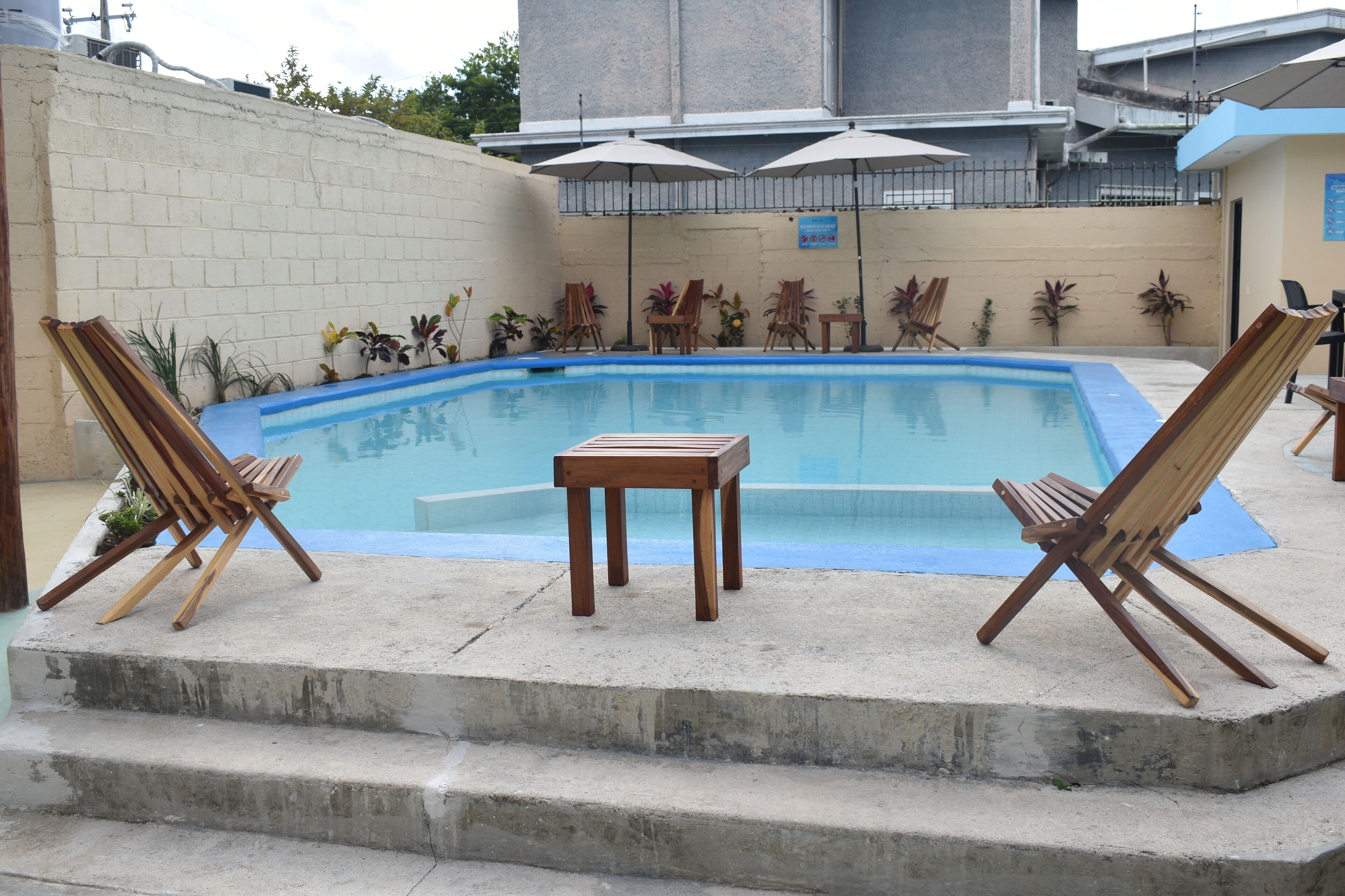 Una piscina con sillas de descanso y una mesita, rodeada de una solana de hormigón y un muro de ladrillo.