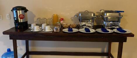 A breakfast setup with a coffee maker, cups, plates, and food containers on a wooden table.