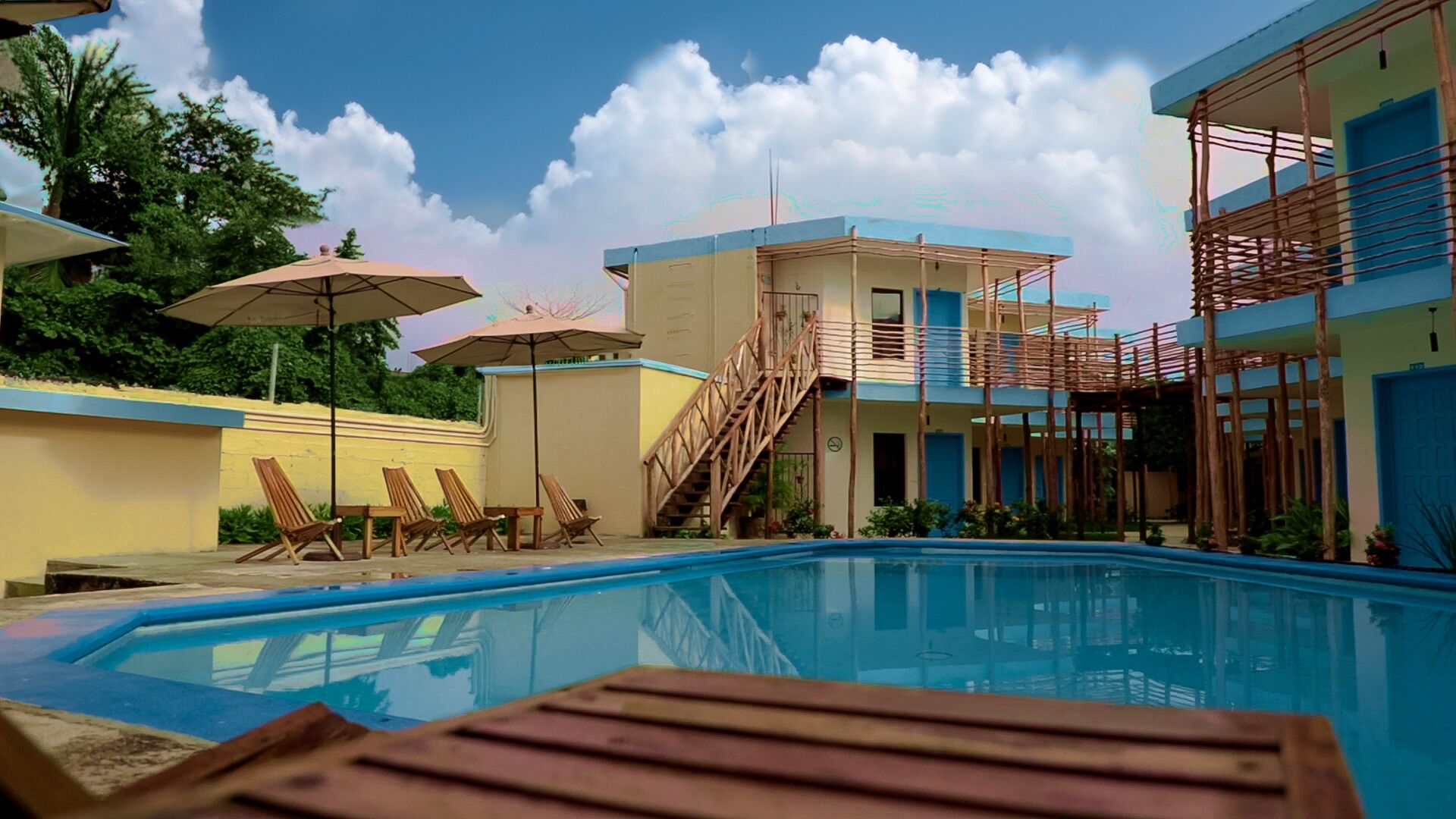 Una piscina con sillones de madera y sombrillas, una escalera que lleva a un edificio y un cielo despejado.