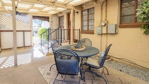A covered patio area with a table and chairs, a small garden, and a building with windows and a door.