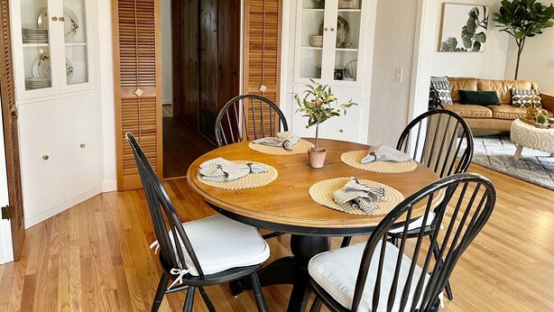 A dining area with a wooden table, black chairs, and a potted plant. There is a clock on the wall and a sofa in the background.