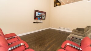 A room with a wooden floor, red chairs, a brown couch, and a mounted screen displaying various streaming services.