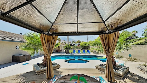 A pool area with a covered seating area, lounge chairs, and a decorative palm tree. - Luxe Retreat - Backyard Oasis (Palm Springs)
