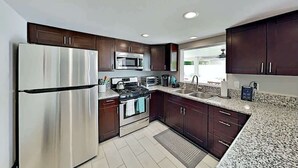 A modern kitchen with stainless steel appliances, dark wooden cabinets, granite countertops, and a large window providing natural light. - Luxe Retreat - Backyard Oasis (Palm Springs)
