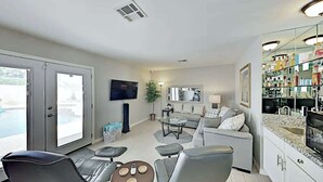 A modern living room with a grey sofa, a coffee table, and a TV. There is a kitchen area with a granite countertop and a refrigerator. - Luxe Retreat - Backyard Oasis (Palm Springs)