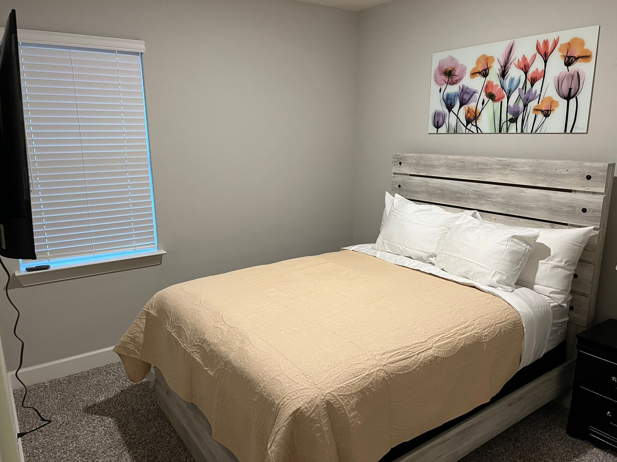A bedroom with a bed, a window with blinds, a painting on the wall, and a nightstand.