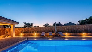 A swimming pool with steps leading into it, surrounded by a stone wall and a covered seating area with chairs and a table.