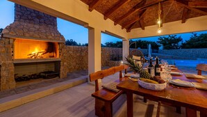 An outdoor dining area with a stone fireplace, wooden furniture, and a pool in the background.