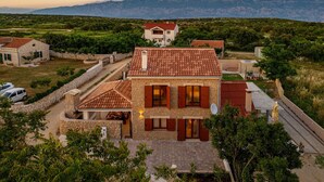 A two-story stone house with a red-tiled roof, surrounded by greenery and a mountainous backdrop.