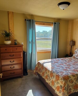 Um quarto com cômoda de madeira, cama com colcha floral, janela com cortinas e vista para um campo gramado.