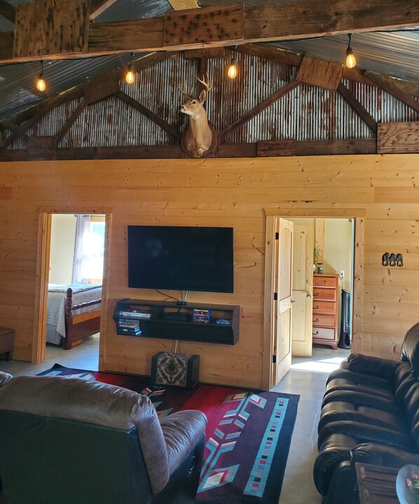 Interior rústico de cabana com teto de madeira, cabeça de veado montada, TV de tela plana, sofá de couro e tapete estampado.
