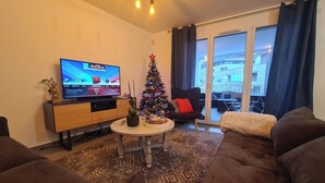 Uma sala de estar com televisão, árvore de Natal, sofá e vista para um edifício pela janela.