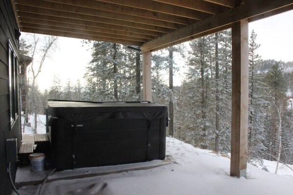 A covered outdoor area with a hot tub, wooden beams, and a snowy forest backdrop.