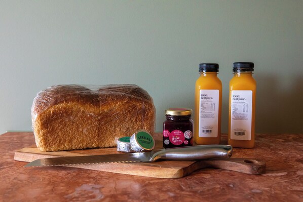 A loaf of bread, two bottles of juice, a jar of jam, and a knife on a cutting board. - The Monty (Anglesea)