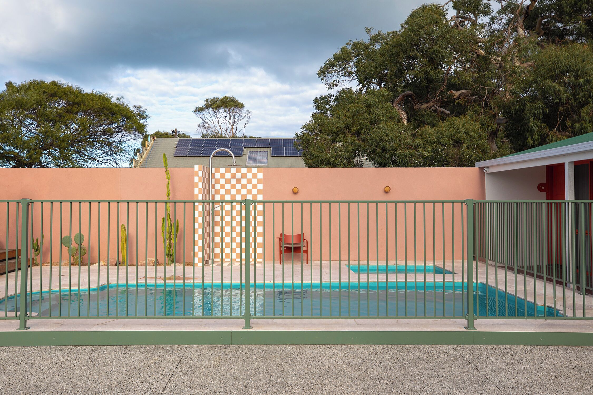 A swimming pool with a fence, a checkered wall, and a building with solar panels in the background.