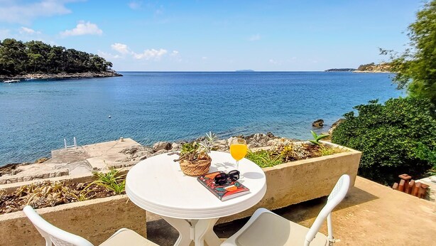 Una mesa con un vaso de jugo de naranja, un libro y gafas de sol, con vista a un mar tranquilo con costas rocosas y vegetación.