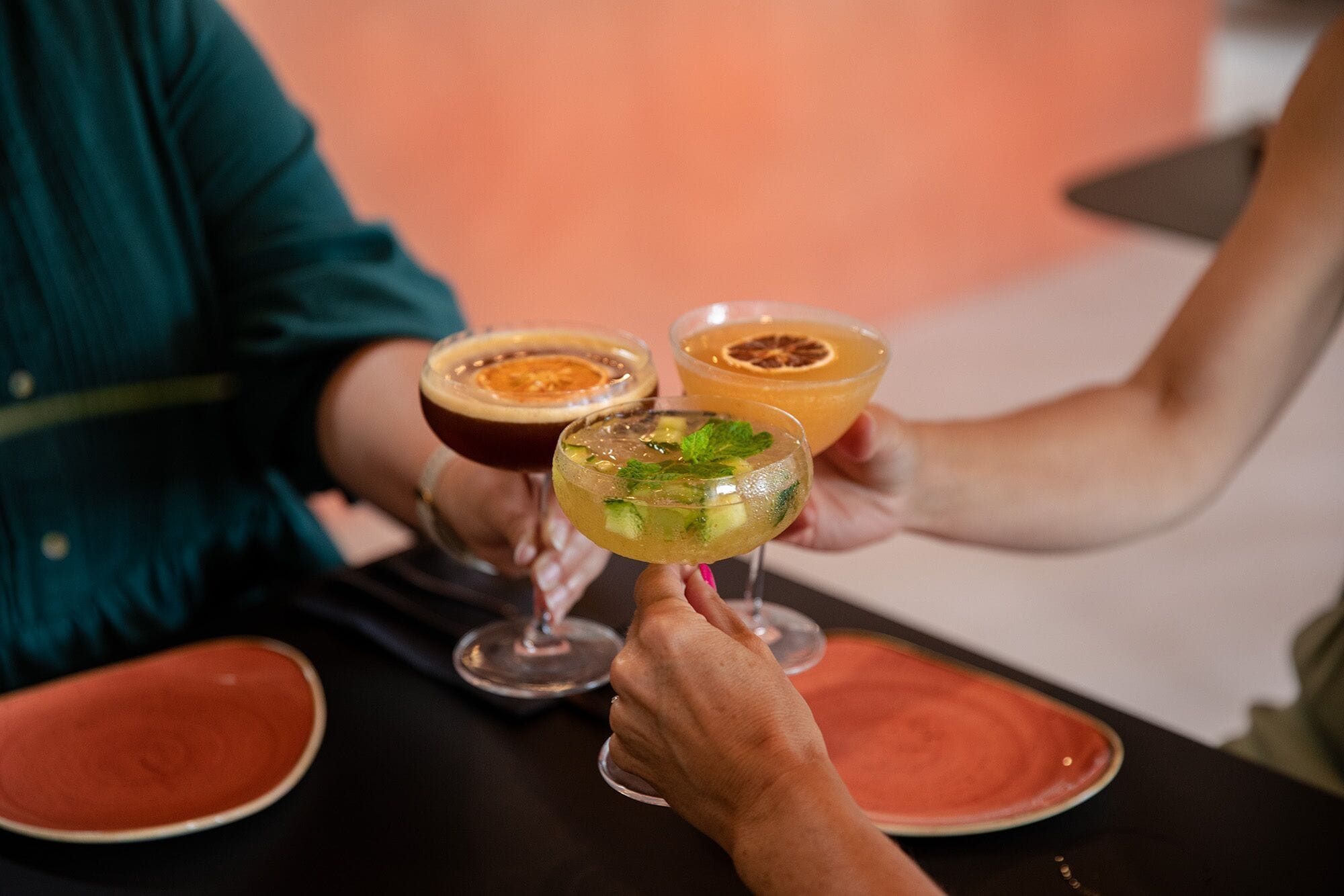 Three cocktails in stemmed glasses, a person holding one, and a table with a red plate.