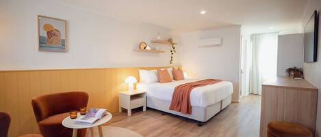 A modern hotel room with a bed, a desk, a chair, a bedside table, and a wall-mounted shelf.