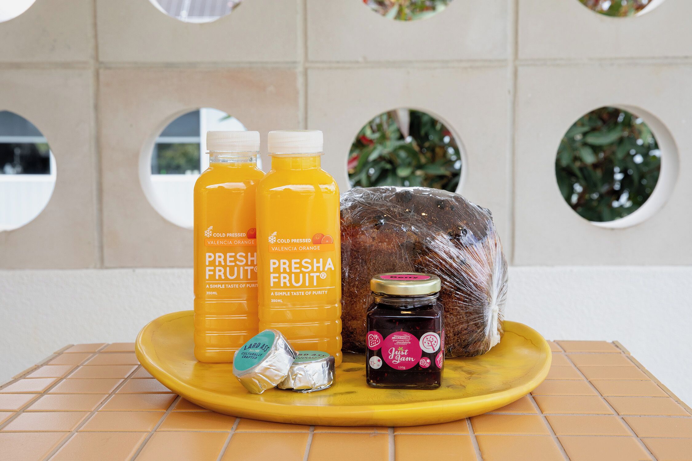 A yellow tray with two bottles of Valencia orange juice, a jar of Presha fruit jam, and a wrapped loaf of bread on a tiled surface.