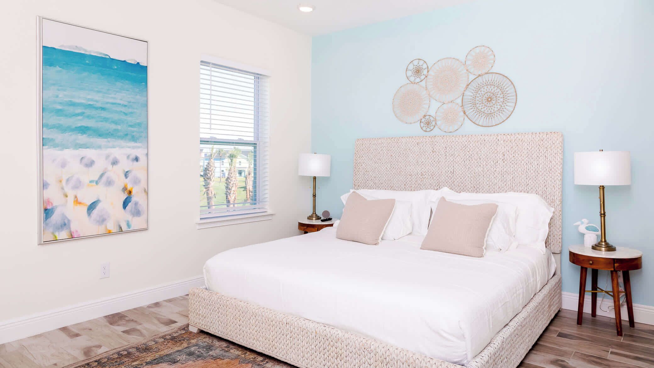 A bedroom with a large bed, two bedside tables with lamps, a window with blinds, a beach-themed painting, and a wall art piece.