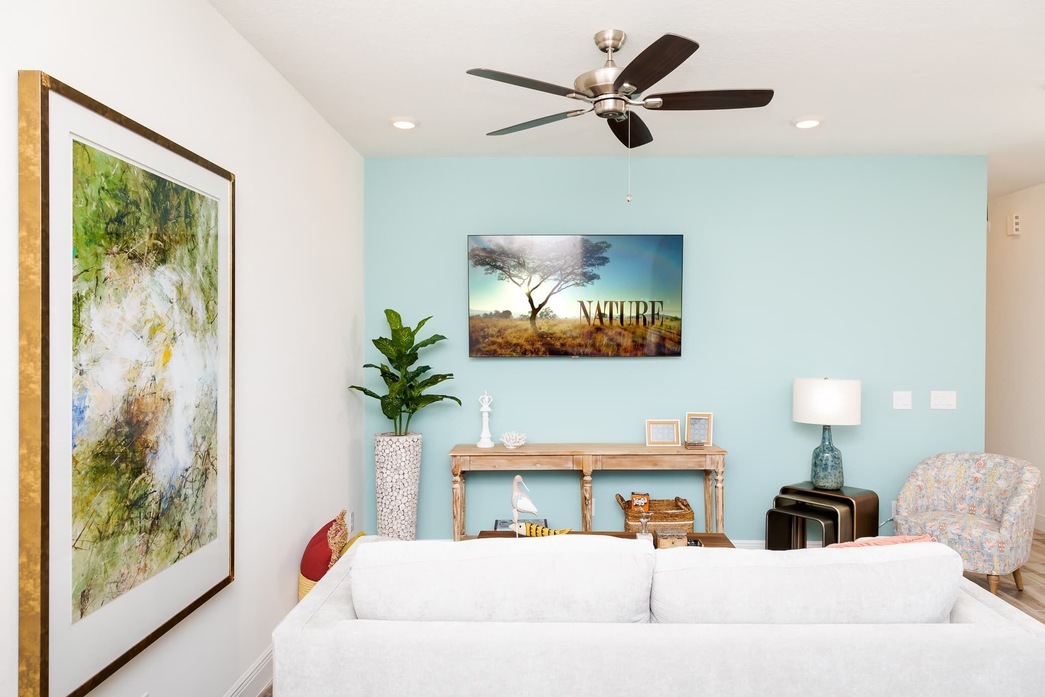 Un salon avec un canapé blanc, un ventilateur de plafond, un tableau représentant un paysage encadré, une console en bois, une lampe et un fauteuil à motifs.