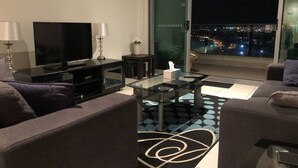 Ett modernt vardagsrum med en stor tv-skärm, ett soffbord i glas och utsikt över staden genom ett stort fönster.