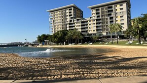 A beachfront hotel with multiple floors, a sandy beach in the foreground, and palm trees.