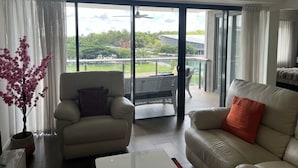 A modern living room with a sofa, armchair, and a small table. There is a balcony with a view of greenery and buildings.