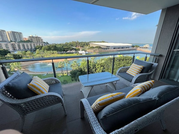 A balcony with wicker furniture, a glass table, and a view of a body of water and buildings.
