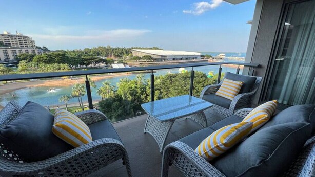 A balcony with wicker furniture and a glass table overlooking a pool and beach.