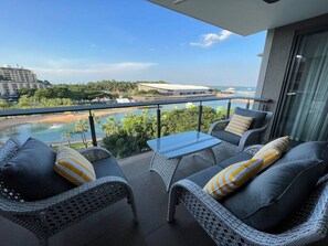 A balcony with wicker furniture and a glass table overlooking a pool and beach.