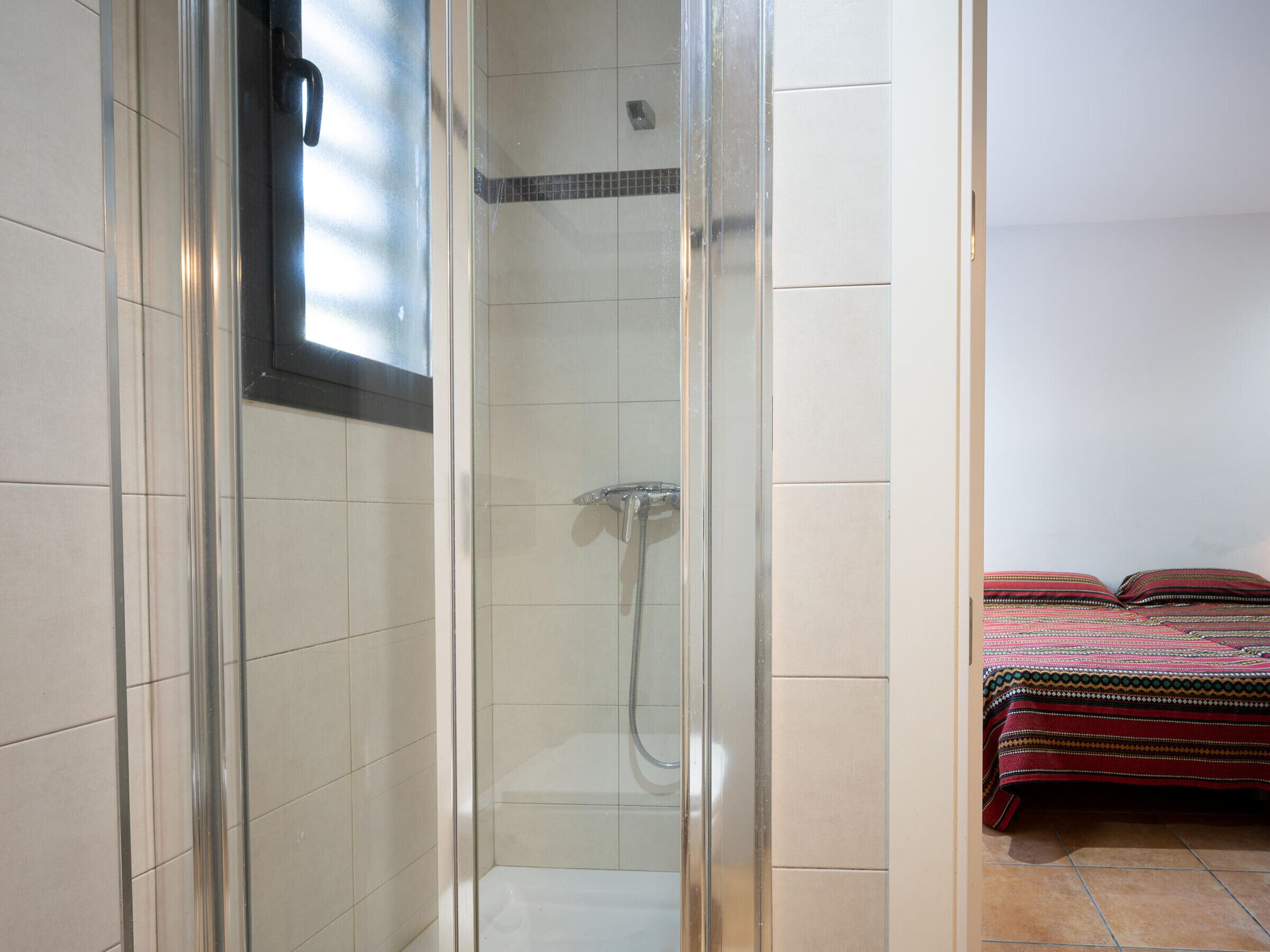 Cabina doccia in vetro con soffione a mano, adiacente a un letto con copripiumino fantasia.