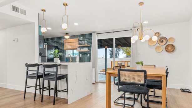 A modern kitchen with a dining area, featuring a wooden table and chairs, a large window, and a hanging light fixture.