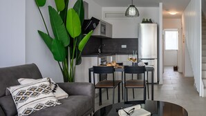 A modern living room with a grey sofa, a coffee table with a book and glasses, and a large green plant. A kitchen is visible in the background with a dining area.
