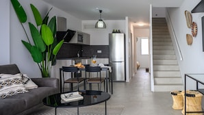 A modern living room with a sofa, coffee table, and chairs. A kitchen with a refrigerator and microwave is visible in the background.