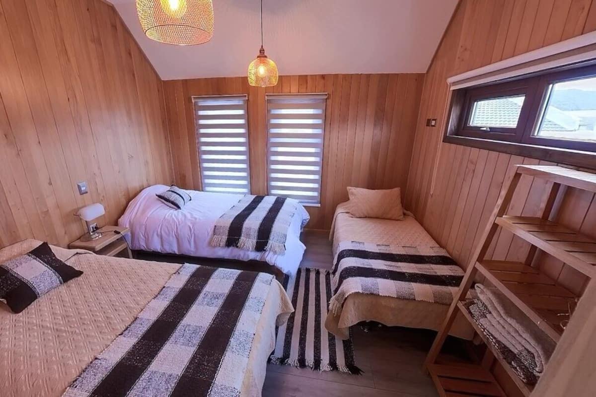 A room with wooden walls, two beds with striped bedding, a nightstand, a lamp, and a window with blinds.