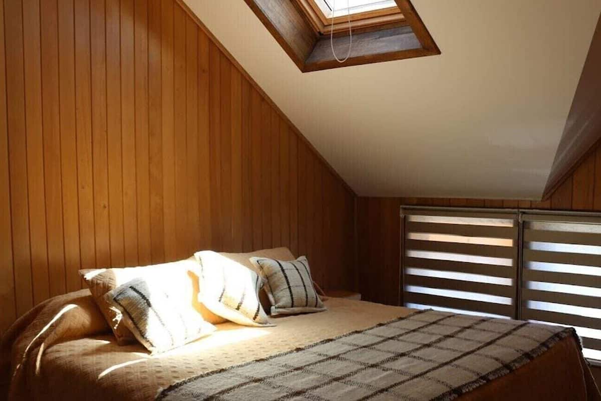 A bedroom with a sloped ceiling, wooden walls, a bed with pillows, a blanket, and a window with blinds.
