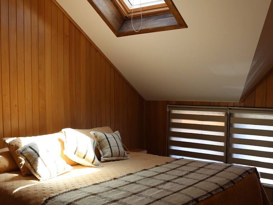 Une chambre avec des lambris en bois, un puits de lumière, un lit avec des oreillers et une fenêtre avec des stores.