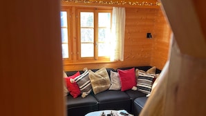 Un accogliente interno di una baita in legno, con un divano nero, un tavolo rotondo in legno e una finestra con tende.
